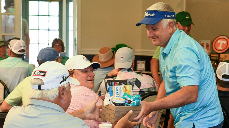 A man in a blue shirt and white visor hands a basket filled with items to another man seated at a table.